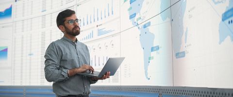 A businessperson holding a laptop stands in front of a projector screen reviewing data.