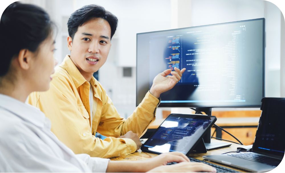 Two people in a work setting are engaged in a discussion as one points at a monitor displaying data 