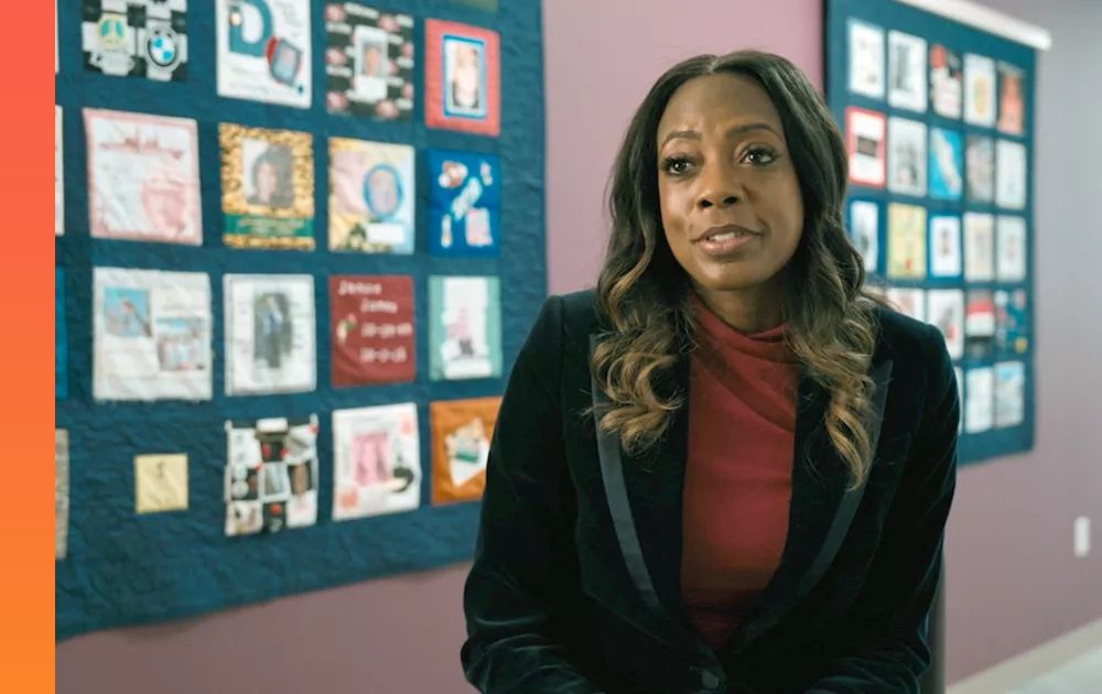 Businessperson in a black blazer and maroon top speaks in front of a wall-mounted quilt featuring various images. 