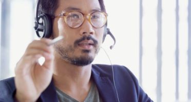 Man in glasses wearing headphones and touching device screen with a play button overlay.